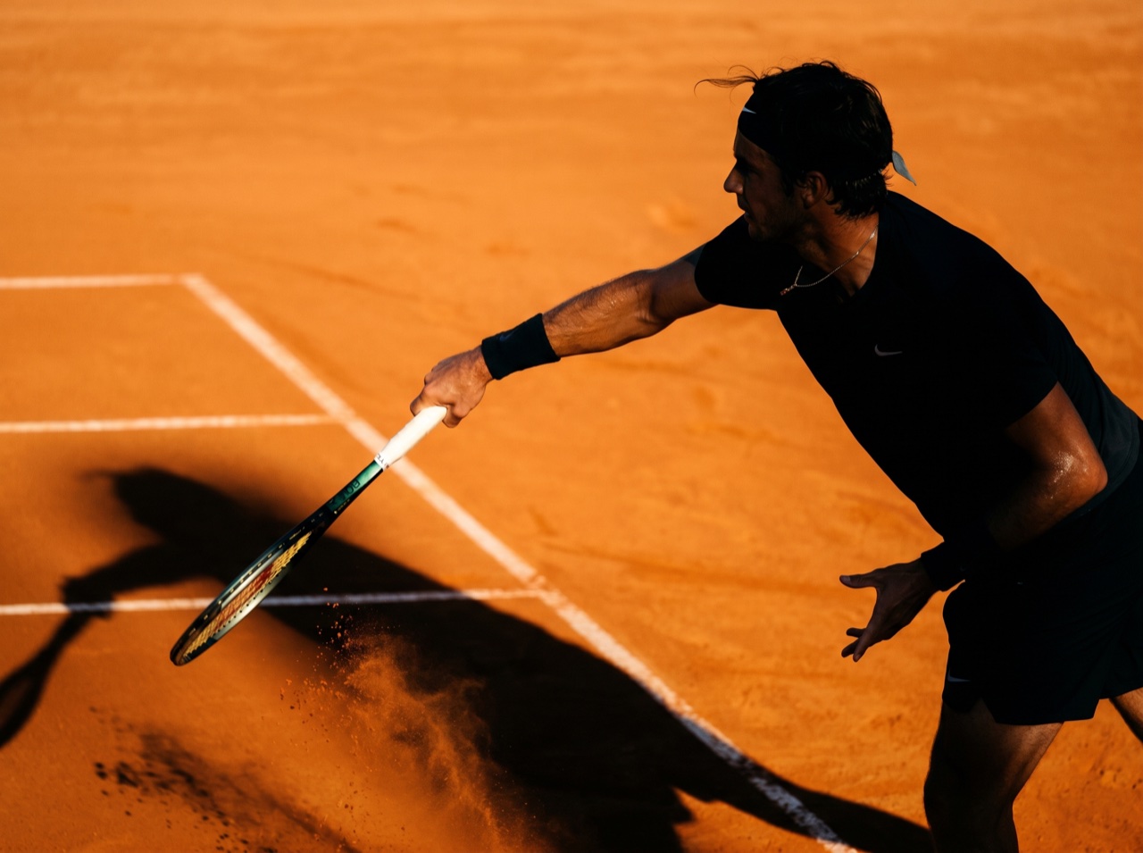 A player mid-serve casting a long shadow on clay