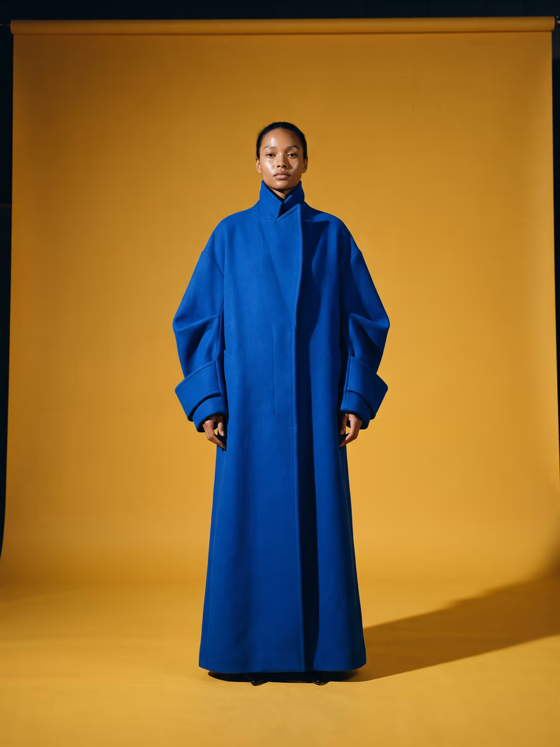 A woman in a cobalt coat on a mustard seamless backdrop, hard side light