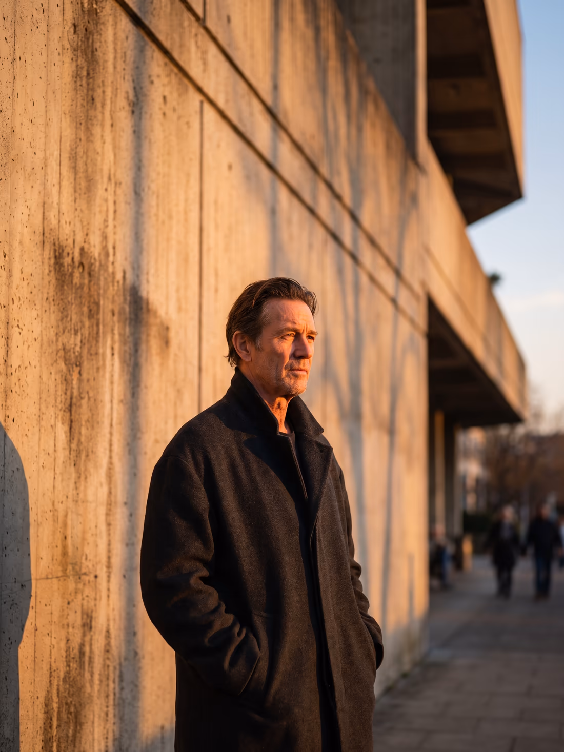 A man in an oversized coat against a brutalist concrete wall at golden hour